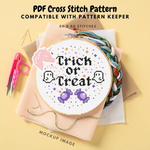 Pode incluir: Um aro de bordado branco com um padrão de ponto cruz que diz "Trick or Treat" em letras pretas. O padrão também inclui duas bengalas de doces roxas, dois fantasmas brancos e um unicórnio rosa com a palavra "Floof" escrita nele. O aro está sobre um tecido rosa com um tecido marrom por baixo.