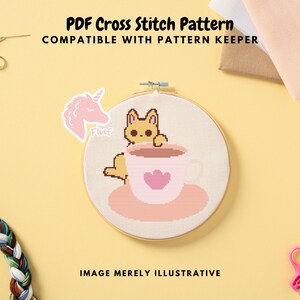 Pode incluir: Um padrão de ponto cruz num bastidor de madeira apresenta um gato de desenho animado numa chávena de café com um coração. O texto "PDF Cross Stitch Pattern" e "Compatible with Pattern Keeper" está no topo. Um autocolante de unicórnio rosa está à esquerda.