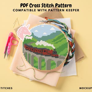 Può includere: Un motivo a punto croce in un telaio di legno raffigura un treno rosso su un ponte, sullo sfondo di un paesaggio verde e cielo blu. L'immagine include forbici rosa, filo da ricamo e il testo "PDF Cross Stitch Pattern".