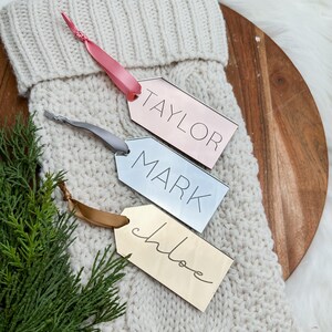 Op de afbeelding: Drie gepersonaliseerde kerstsoklabels met de namen "Taylor", "Mark" en "Chloe" geschreven in gouden, zilveren en roze letters. De labels zijn bevestigd aan een witte gebreide sok.