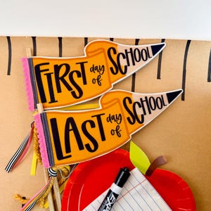 Könnte beinhalten: Zwei Wimpel in Stiftform mit orangefarbenen Körpern und schwarzem Text mit der Aufschrift First Day of School und Last Day of School. Die Wimpel haben rosa Bommelborten, Holzstäbe und Bänder. Ein roter Teller, ein Marker und Papier sind dabei.