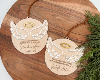 Op de afbeelding: Twee houten engelenvleugel-versieringen met een gouden halo. De ornamenten hebben de tekst "Je vleugels waren klaar, maar onze harten niet" en de namen "Grandma Anne 1938-2023" en "Wade John".