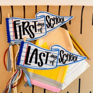 Könnte beinhalten: Zwei Filzwimpel an Holzstäben mit blau-weißem Notizbuchmuster. Einer trägt die Aufschrift First Day of School, der andere Last Day of School in schwarzer Schrift. Sie haben blaue Bommelborten und Bänder sowie einen bleistiftförmigen Pappteller.