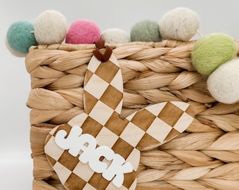 May include: A wooden bunny-shaped Easter basket tag with a checkered pattern in brown and white. The tag has the name "Jack" written in white letters.