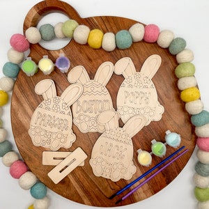 Può includere: Quattro forme di uova di Pasqua di coniglio in legno non finite con i nomi Parker, Boston, Poppy e Jack. Le forme sono su una superficie di legno con barattoli di vernice e pennelli.