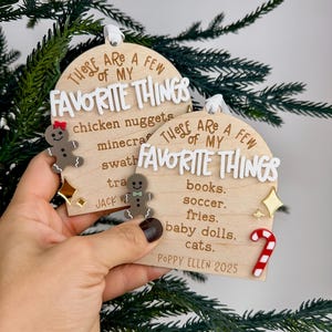 May include: Two wooden Christmas ornaments with the text "These are a few of my favourite things." One ornament has a gingerbread man and the other has a candy cane. The ornaments are held in front of a green Christmas tree.