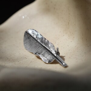 May include: A silver feather-shaped brooch with a detailed design. The brooch is on a light beige surface.