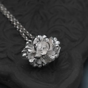 Op de afbeelding: Een zilverkleurige hangerketting met een gedetailleerd bloemdesign. De hanger is een driedimensionale bloem met ingewikkelde bloemblaadjes. De ketting is een delicaat, geschakeld ontwerp.