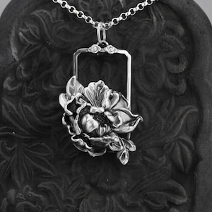 May include: A silver pendant necklace featuring a detailed floral design. The flower is a peony with a delicate frame around it. The necklace is on a black background.