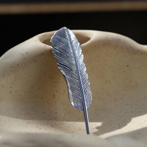 May include: A silver feather-shaped brooch with a detailed design. The brooch is sitting on a light brown textured surface.