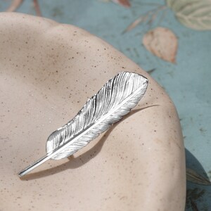 May include: A silver feather-shaped brooch with a detailed design. The brooch is lying on a white surface.