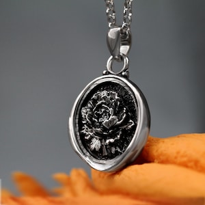 May include: A silver pendant necklace with a round, black, wax seal-like charm featuring a detailed, embossed image of a flower.