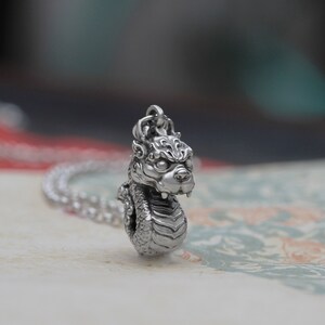 May include: A silver pendant necklace featuring a detailed dragon head design. The dragon has a fierce expression and is coiled in a defensive posture.