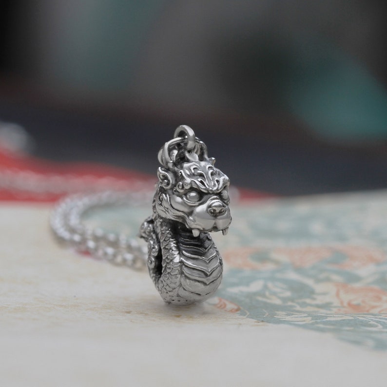 May include: A silver pendant necklace featuring a detailed dragon head design. The dragon has a fierce expression and is coiled in a defensive posture.