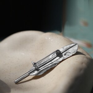 May include: A silver feather-shaped brooch with a detailed design. The brooch has a pin on the back for attaching to clothing.
