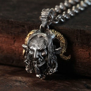 May include: Silver and gold pendant necklace featuring a detailed sculpted head of a horned creature with a human-like face. The pendant is attached to a silver chain.