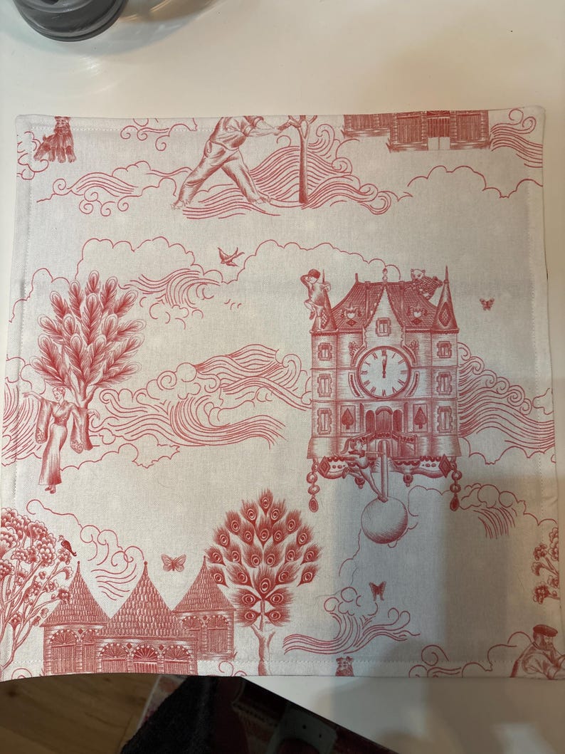 K&ouml;nnte beinhalten: Ein quadratischer, cremefarbener Tischset mit rotem Toile-Muster. Das Design umfasst Illustrationen eines Uhrturms, B&auml;ume, Figuren und Wolken. Das Tischset hat einen gen&auml;hten Rand.