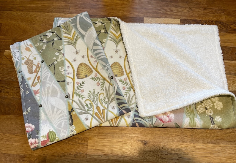 Peut inclure: Une couverture r&eacute;versible pour b&eacute;b&eacute; avec un motif botanique. Un c&ocirc;t&eacute; pr&eacute;sente un patchwork de tissus &agrave; motifs floraux et naturels avec des abeilles, des &eacute;cureuils et des fleurs. L'autre c&ocirc;t&eacute; est en mati&egrave;re pelucheuse blanche et douce. La couverture est pli&eacute;e, r&eacute;v&eacute;lant les deux c&ocirc;t&eacute;s.