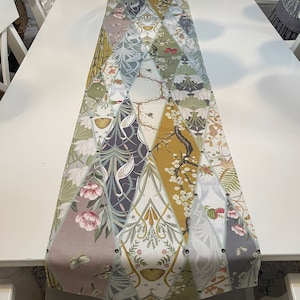 Può includere: Runner da tavolo decorativo con un design patchwork con motivi floreali e di uccelli nei toni del verde, rosa, oro e grigio. Il runner è posto su un tavolo bianco e presenta borchie metalliche sui bordi. Misura circa 150 cm.