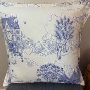 K&ouml;nnte beinhalten: Quadratisches Kissen mit blau-wei&szlig;em Toile-de-Jouy-Design. Das Kissen zeigt eine detaillierte Illustration eines Uhrturms, Figuren, B&auml;ume und Wolken. Das Kissen ist aus einem weichen Stoff gefertigt.