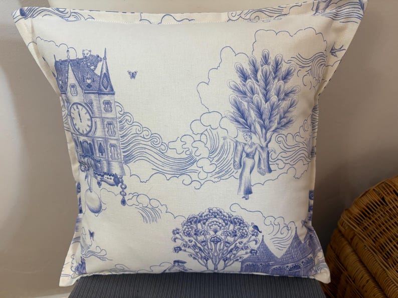 K&ouml;nnte beinhalten: Quadratisches Kissen mit blau-wei&szlig;em Toile-de-Jouy-Design. Das Kissen zeigt eine detaillierte Illustration eines Uhrturms, Figuren, B&auml;ume und Wolken. Das Kissen ist aus einem weichen Stoff gefertigt.