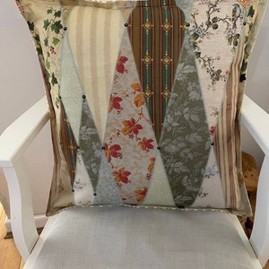 May include: Square decorative pillow with a patchwork design. The pillow features various fabric patterns, including floral, striped, and leaf motifs in beige, green, and brown tones. Small black studs accent the seams.