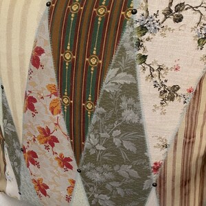 Peut inclure: Gros plan d'un coussin d&eacute;coratif avec un motif patchwork. Le coussin pr&eacute;sente divers tissus &agrave; motifs, notamment des rayures, des imprim&eacute;s floraux et des motifs de feuilles dans des tons de beige, marron, vert et rouge. Des clous en m&eacute;tal fonc&eacute; sont plac&eacute;s le long des coutures.