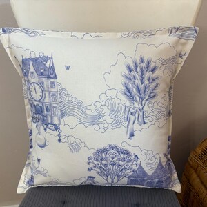 K&ouml;nnte beinhalten: Quadratisches Zierkissen mit blau-wei&szlig;em Toile-de-Jouy-Muster. Das Design zeigt einen Uhrturm, Figuren, B&auml;ume und Wolken. Das Kissen liegt auf einer grauen Oberfl&auml;che.