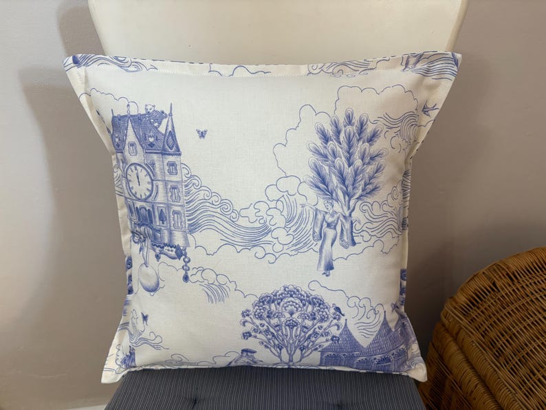 K&ouml;nnte beinhalten: Quadratisches Zierkissen mit blau-wei&szlig;em Toile-de-Jouy-Muster. Das Design zeigt einen Uhrturm, Figuren, B&auml;ume und Wolken. Das Kissen liegt auf einer grauen Oberfl&auml;che.