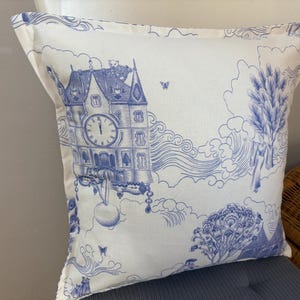 K&ouml;nnte beinhalten: Quadratisches Zierkissen mit blau-wei&szlig;em Toile-de-Jouy-Design. Das Kissen zeigt eine detaillierte Illustration eines Uhrturms, Wolken und anderer dekorativer Elemente. Das Kissen liegt auf einem grauen Stuhl.
