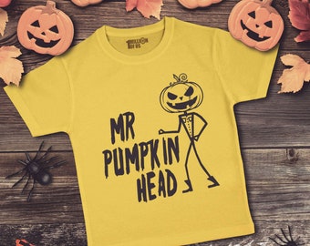 May include: A yellow t-shirt with a black pumpkin head graphic and the text "Mr. Pumpkin Head". The shirt is on a wooden background with pumpkins, leaves, and spiders.