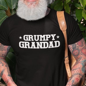 Op de afbeelding: Een zwart T-shirt met witte tekst die "*GRUMPY* GRANDAD" zegt. De persoon die het shirt draagt heeft een witte baard en lacht.