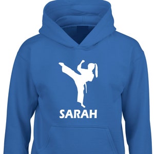 Puede incluir: Una sudadera con capucha de color azul brillante que presenta una silueta gr&aacute;fica blanca de una persona realizando una patada de artes marciales. Debajo del gr&aacute;fico, el nombre SARAH est&aacute; impreso en letras may&uacute;sculas blancas. Bolsillo frontal.
