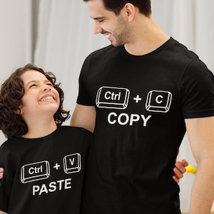 May include: Black t-shirts with white text. One shirt says "Ctrl + C COPY" and the other says "Ctrl + V PASTE". The shirts are worn by a person and a child, who are smiling at each other.