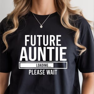 May include: Black t-shirt with the text "FUTURE AUNTIE" in large white letters. Below, a loading bar graphic with "LOADING... PLEASE WAIT" in white. The shirt is worn by a person with blonde hair and a silver necklace.