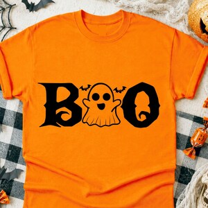 Può includere: Una t-shirt a maniche corte color arancione brillante con la parola BOO stampata in nero. La lettera O centrale è sostituita dal disegno di un fantasma sorridente con due piccoli pipistrelli sopra la testa. Il testo ha un carattere stilizzato.