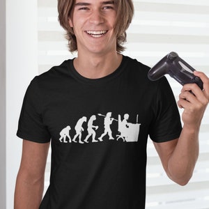 May include: A black t-shirt featuring a white graphic depicting the evolution of man, culminating in a person seated at a computer. The person is holding a controller in their right hand.