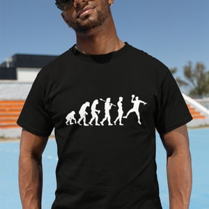 Può includere: T-shirt nero con grafica bianca raffigurante l'evoluzione di un giocatore di pallamano. Il design mostra una progressione da figure simili a scimmie a una persona che lancia una palla da pallamano. La maglietta è indossata da una persona con occhiali da sole.