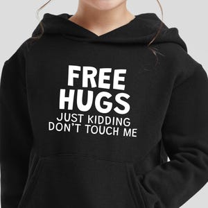 以下が含まれることがあります： 白文字で「FREE HUGS JUST KIDDING DON'T TOUCH ME」と書かれた黒いパーカー。 パーカーにはフロントポケットとドローストリングフードが付いています。