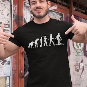 Könnte beinhalten: Ein schwarzes T-Shirt mit einer weißen Grafik der Evolution des Menschen, die mit einem Skifahrer endet. Die Grafik zeigt eine Progression von einem primitiven Affen zu einem modernen Menschen, die mit einem Skifahrer auf Skiern gipfelt.