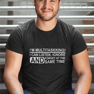 May include: Black t-shirt with white text: "I'M MULTITASKKING! I CAN LISTEN, IGNORE AND FORGET AT THE SAME TIME." A person is wearing the shirt while sitting on steps.
