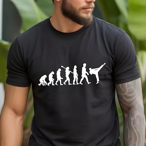 May include: A black short-sleeved t-shirt featuring a white graphic print on the chest. The design depicts the evolution of humanity, starting with a primate and progressing through various stages to a figure performing a high martial arts kick.