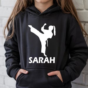 Puede incluir: Una sudadera con capucha negra que presenta una silueta gr&aacute;fica blanca de una persona realizando una patada alta con uniforme de artes marciales. Debajo del gr&aacute;fico, el nombre SARAH est&aacute; impreso en letras may&uacute;sculas blancas. Bolsillo frontal.