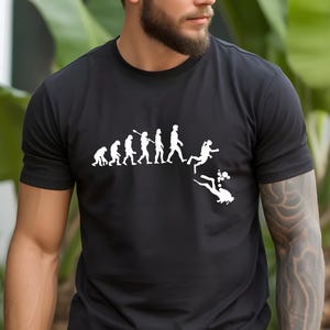 May include: A black short-sleeved t-shirt featuring a white graphic print that depicts the human evolution sequence, transitioning from a primate to a standing human, and finally into a scuba diver swimming underwater with rising air bubbles.