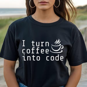 May include: A black short-sleeved t-shirt featuring white text that reads I turn coffee into code, accompanied by a graphic of a steaming coffee cup. The shirt has a crew neckline and is worn with blue denim jeans, visible at the waistline.