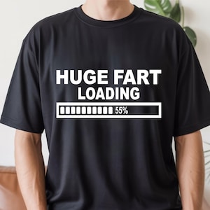 May include: A black short-sleeved t-shirt featuring white text that reads HUGE FART LOADING above a progress bar graphic with the text 55%. The design is centred on the chest of the garment, which is worn with blue denim jeans and a brown belt.