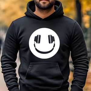 Op de afbeelding: Een zwarte hoodie met lange mouwen en een grote witte cirkelvormige afbeelding op de borst. De afbeelding toont een gestileerd smileygezicht met een koptelefoon. De trui is voorzien van een kangoeroezak en geribbelde boorden aan de mouwen.