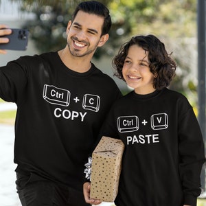 May include: Two people wearing black sweatshirts with white text. One sweatshirt says "Ctrl + C COPY" and the other says "Ctrl + V PASTE". They are holding a phone and a wrapped gift.