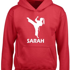 Puede incluir: Una sudadera con capucha de color rojo brillante que presenta un gr&aacute;fico blanco de un artista marcial realizando una patada alta. Debajo del gr&aacute;fico, el nombre SARAH est&aacute; impreso en letras may&uacute;sculas blancas. La prenda incluye mangas largas y un bolsillo.