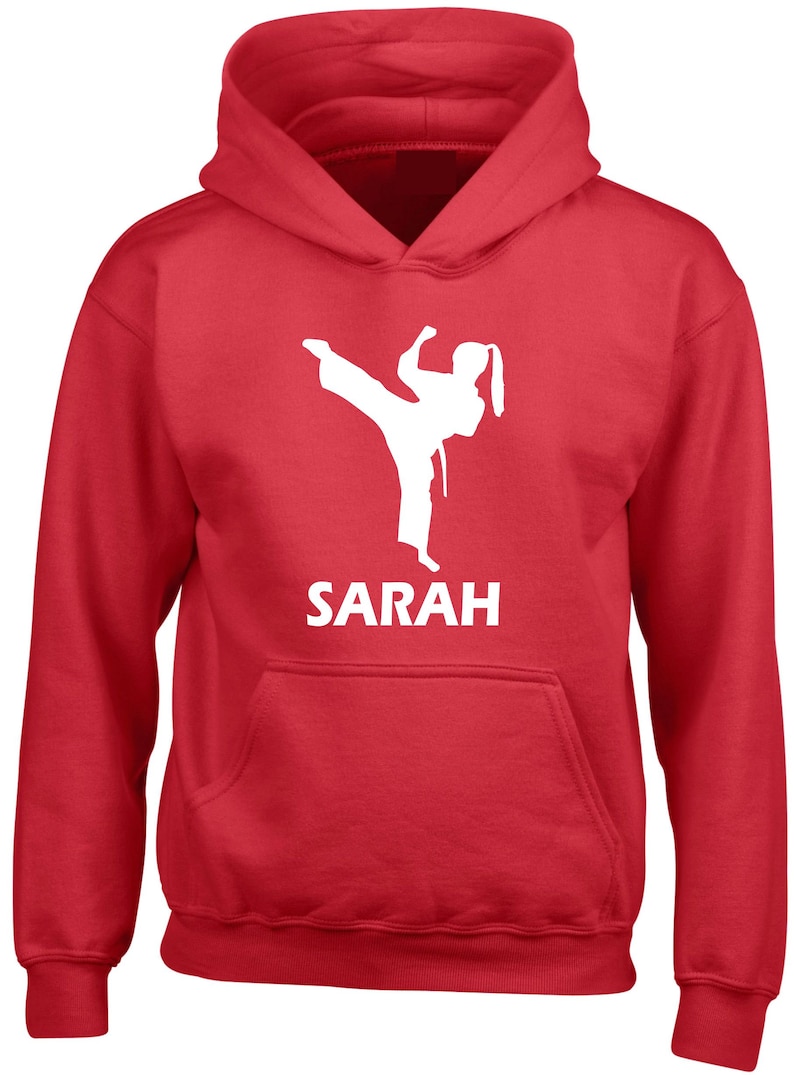 Puede incluir: Una sudadera con capucha de color rojo brillante que presenta un gr&aacute;fico blanco de un artista marcial realizando una patada alta. Debajo del gr&aacute;fico, el nombre SARAH est&aacute; impreso en letras may&uacute;sculas blancas. La prenda incluye mangas largas y un bolsillo.